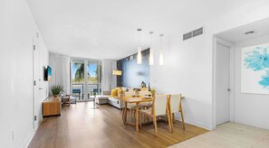 A modern living room with a dining area, large windows, and a view of palm trees. - 2BR • Marina Views at Beachwalk (Hallandale Beach)