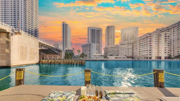 A waterfront view with high-rise buildings in the background, a dock with caution signs, and a table with food and drinks. - Contemporary 1BR Stay | Beach Area + Resort Life (Hallandale Beach)