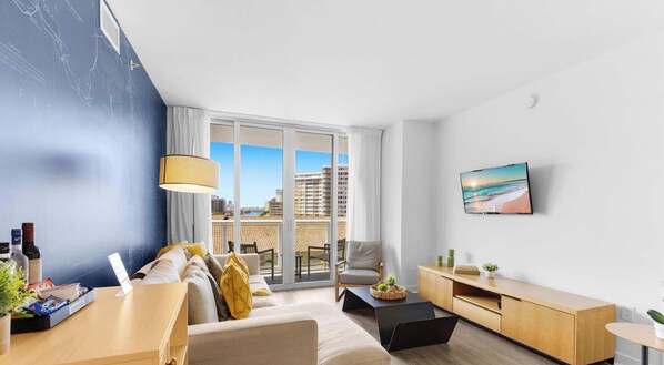 A modern living room with a sofa, coffee table, and TV, featuring a city view through large windows. - 2BR • Intracoastal Views, Hallandale (Hallandale Beach)