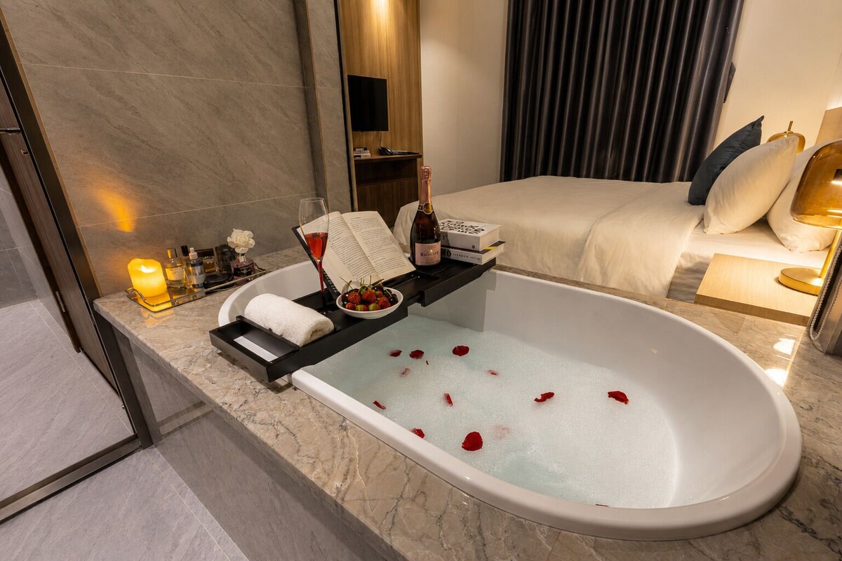 A modern bathroom with a bathtub, a vanity with toiletries, a bed, and a large mirror.