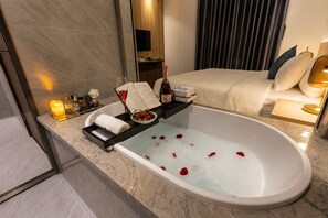 Une salle de bain moderne avec une baignoire, un meuble-lavabo avec des articles de toilette, un lit et un grand miroir.
