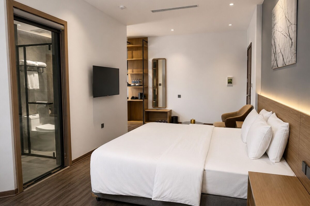 A modern hotel room with a large bed, a television, a seating area, and a bathroom with a glass door.