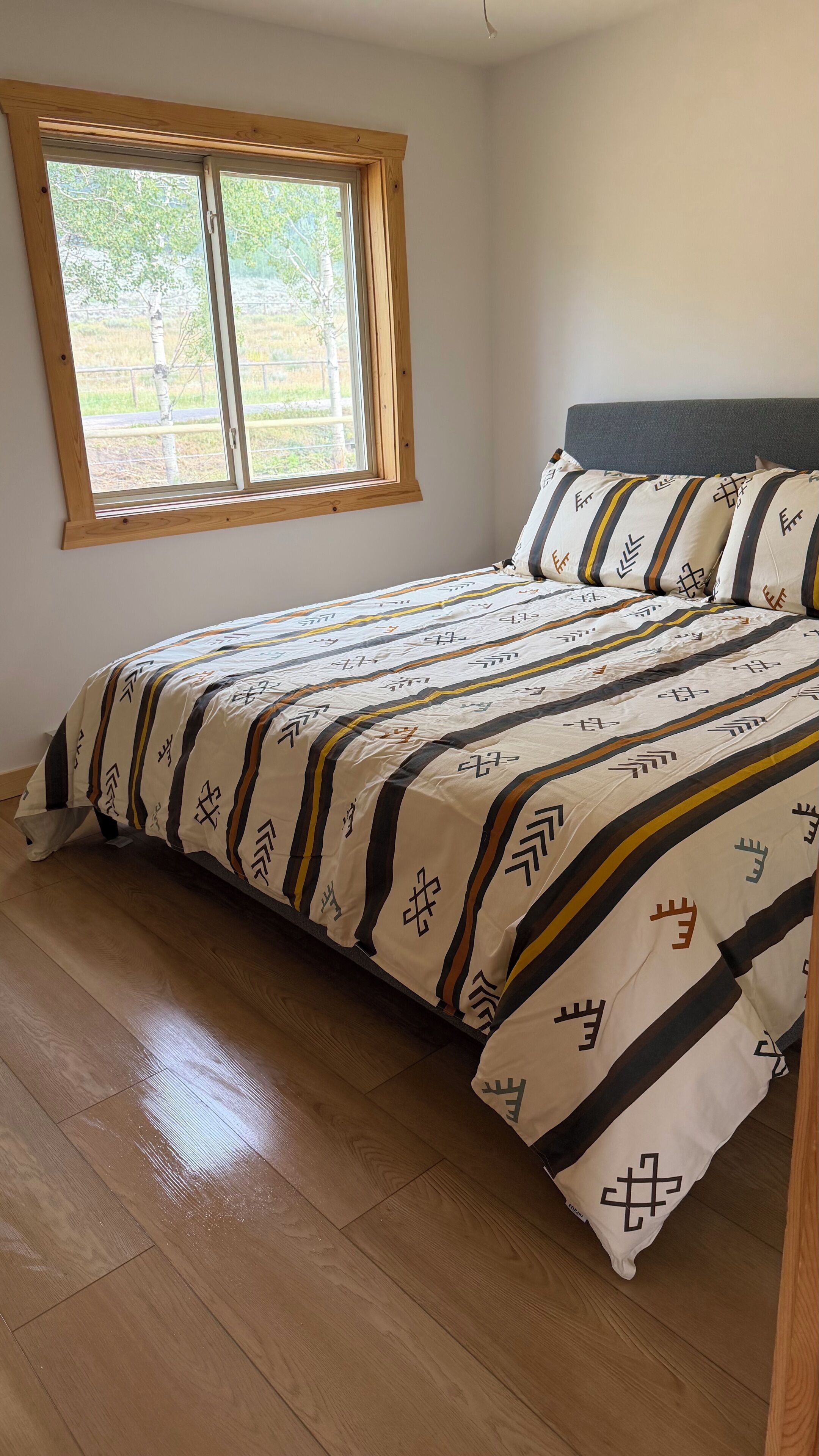 Une chambre avec un plancher en bois, un lit avec une literie à motifs, une fenêtre donnant sur un paysage verdoyant et une tête de lit en bois.