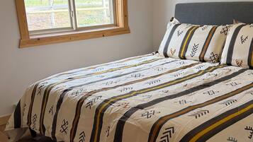 Une chambre avec un plancher en bois, un lit avec une literie à motifs, une fenêtre donnant sur un paysage verdoyant et une tête de lit en bois.