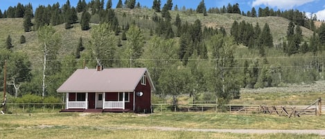 Una casa de un piso con porche, rodeada por un campo cubierto de hierba y una colina boscosa.