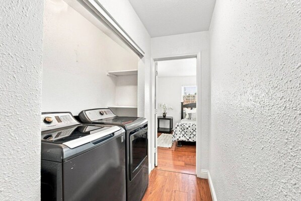 A laundry room with two washing machines and a dryer, leading to a bedroom with a bed and a nightstand.