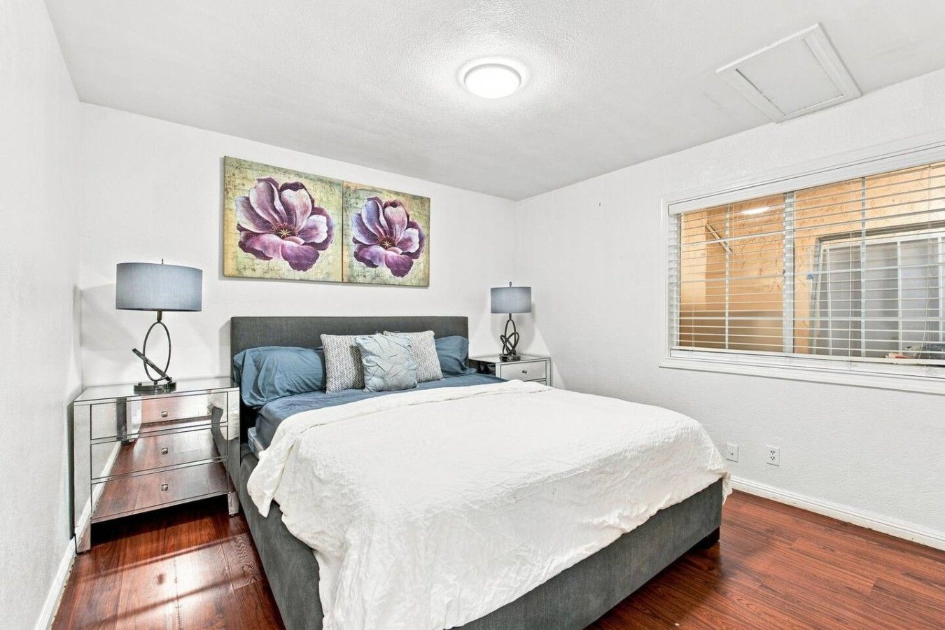 Une chambre à coucher comprenant un lit, deux tables de chevet, une fenêtre avec des stores et un luminaire au plafond.
