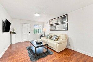 A living room with a beige sofa, a coffee table, a flat-screen TV mounted on the wall, and two framed artworks above the sofa.