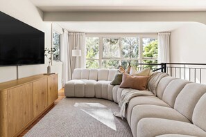 A modern living room with a sectional sofa, a flat-screen TV mounted on the wall, and a large window with curtains.
