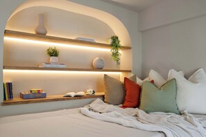 A bedroom with a bed, pillows, a blanket, a shelf with books, a vase, and a plant.