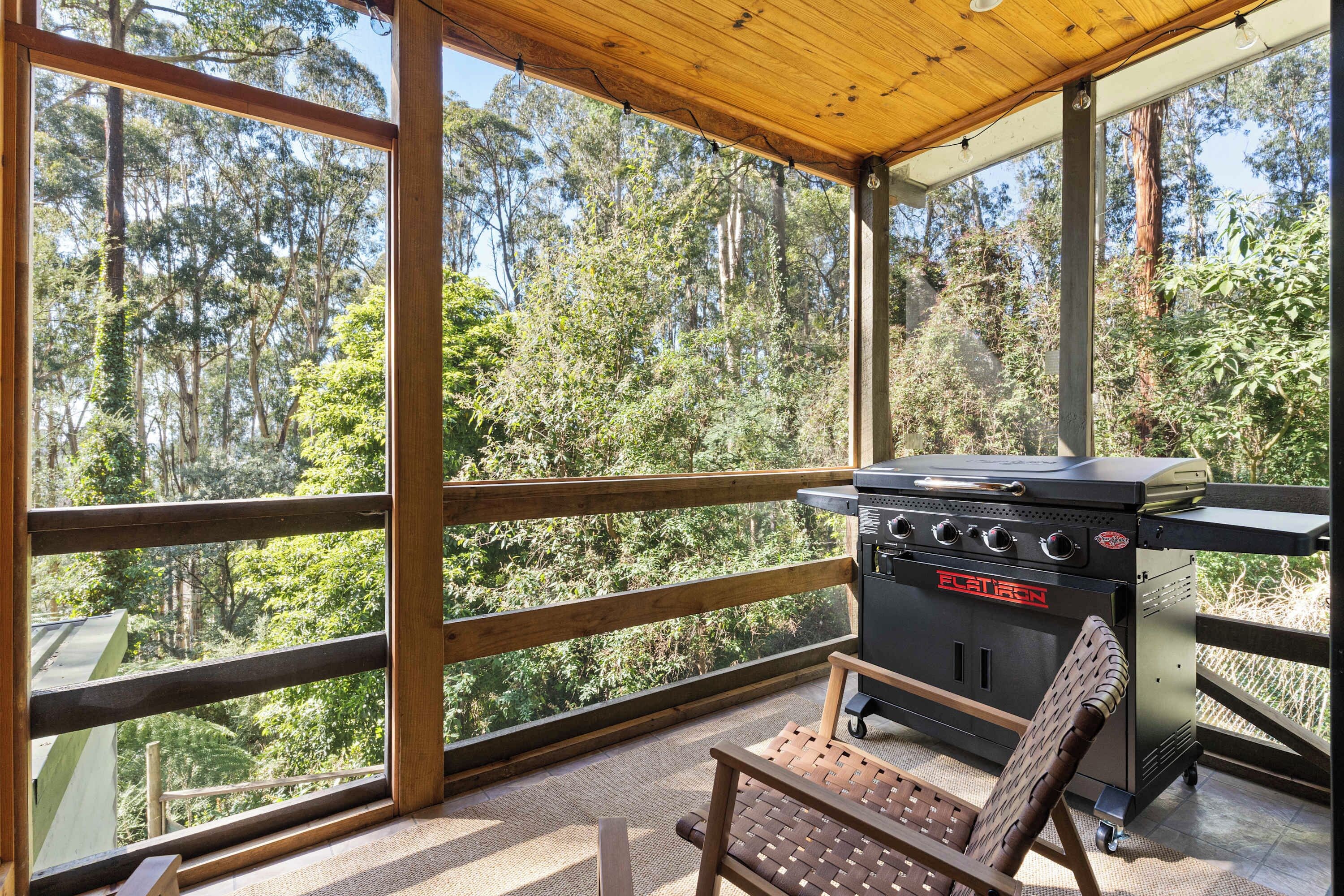 A covered outdoor area with a grill and seating, surrounded by a forest view.