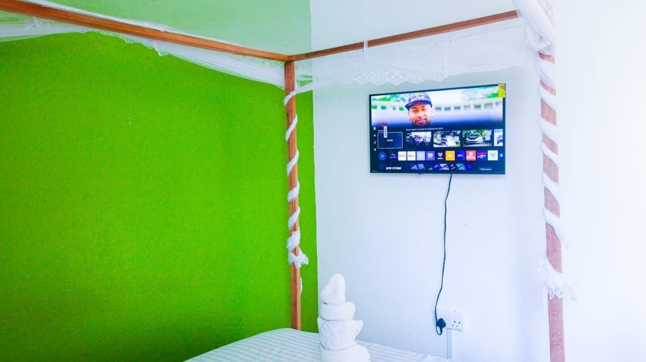 Quarto com parede verde, cama com roupa de cama branca, televisão montada e uma tomada.