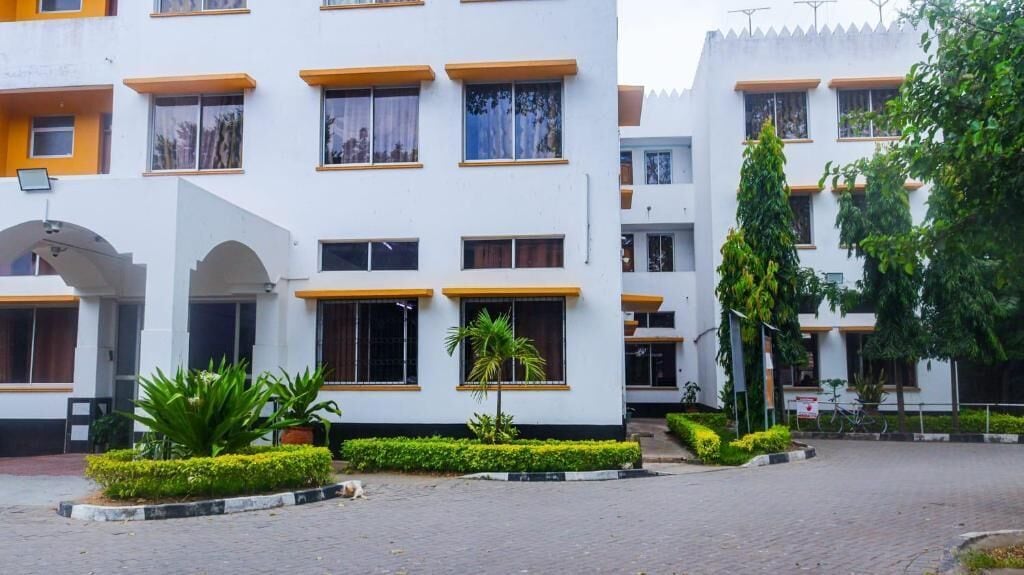 A multi-story building with a white facade and yellow window frames. There are multiple windows, some with curtains, and a small garden area with trimmed bushes and a palm tree.