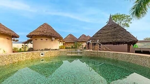 A clear swimming pool surrounded by thatched-roof structures. - forest mud villas (Haridwar)