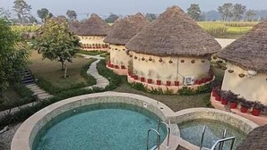 A pool area with a circular hot tub, surrounded by thatched-roof huts, and a pathway leading to them. - forest mud villas (Haridwar)