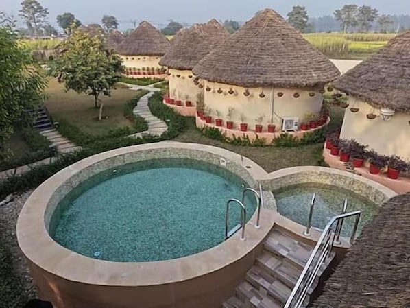 Un área de piscina con un jacuzzi circular, rodeado de cabañas con techo de paja, y un camino que conduce a ellas.