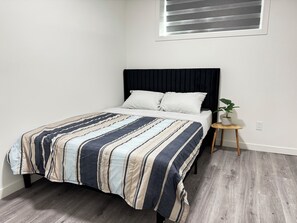 Een bed met een gestreept beddegoed, een nachtkastje met een plant, en een raam met luiken.
