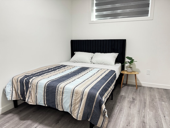 A bed with a striped bedspread, a bedside table with a plant, and a window with blinds.