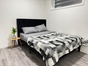 Een bed met een zwart hoofdeinde, grijs beddengoed en een geruit plaid. Er staat een klein houten nachtkastje met een potplant naast het bed.