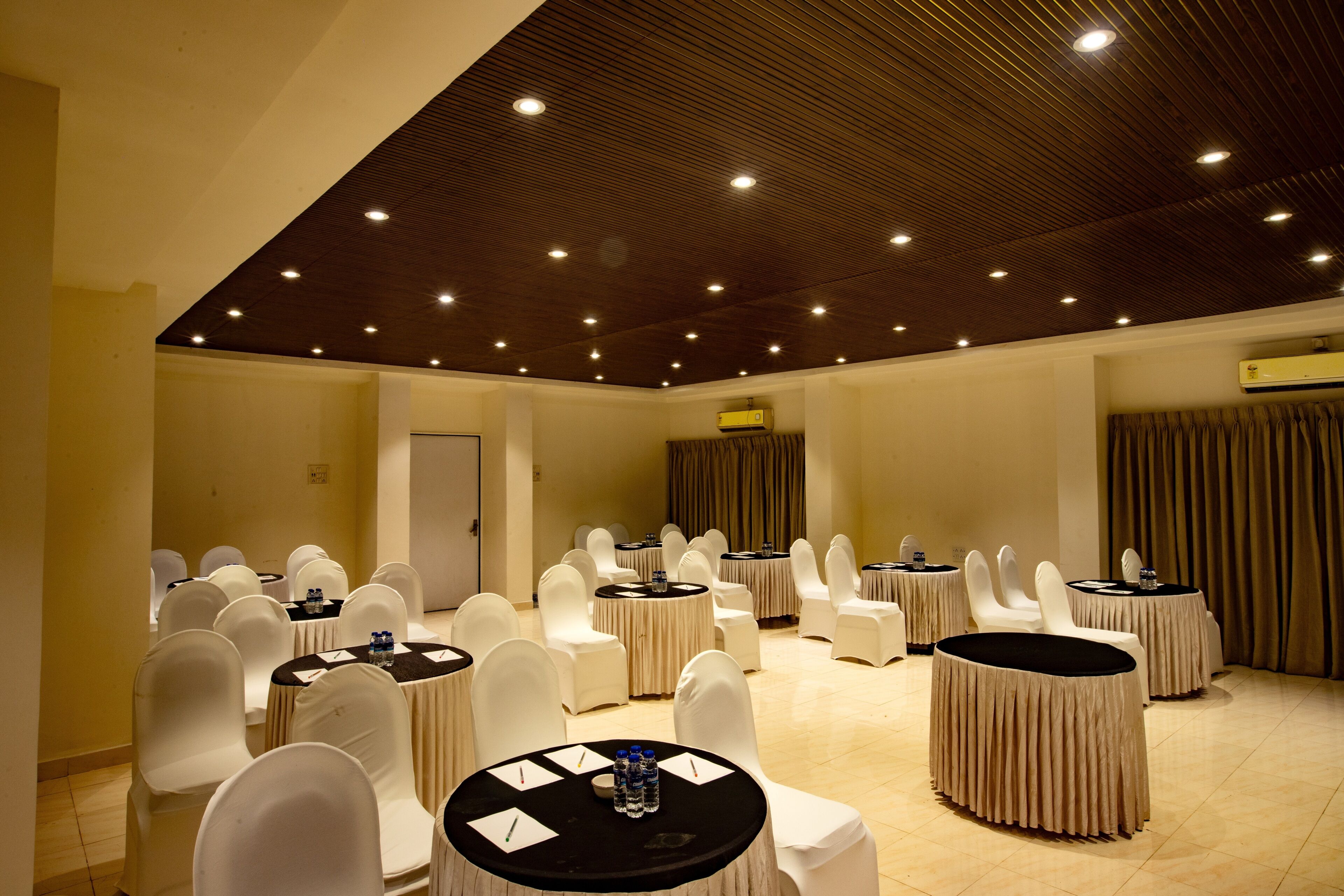 Un salón de conferencias con mesas redondas, sillas blancas y techo de madera con iluminación empotrada.