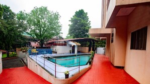 En swimmingpool med ett rött, kakelbeklätt poolområde, omgiven av en byggnad med ett överbyggt område och krukväxter.