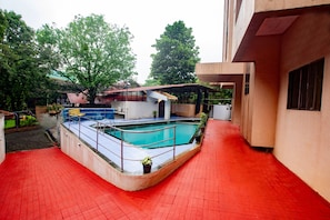 Une piscine dotée d’une terrasse carrelée de carreaux rouges, entourée d’un bâtiment comprenant une zone abritée et des plantes en pot.