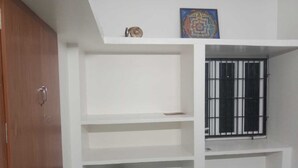 Quarto com guarda-roupa de madeira, cama com colcha estampada, janela com grades de metal e prateleira com objeto decorativo.