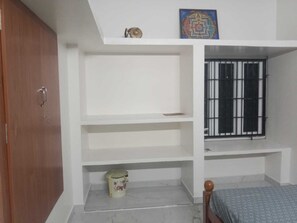 A bedroom with a wooden wardrobe, a bed with a patterned bedspread, a window with metal bars, and a shelf with a decorative item. - Mangai Homestay — A Quiet, Nature-Led Stay in Tiruchendur (Thiruchendur)