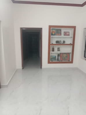 A hallway with a marble floor leading to a door, a wall-mounted shelf with various items, and a plain white wall.