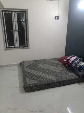 A room with a bed, a window with bars, a wall-mounted light, and a tiled floor. - Mangai Homestay — A Quiet, Nature-Led Stay in Tiruchendur (Thiruchendur)