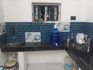 A kitchen with a black countertop, a blue water dispenser, a sink, and a window with bars. - Mangai Homestay — A Quiet, Nature-Led Stay in Tiruchendur (Thiruchendur)
