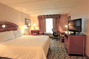 Ein Hotelzimmer mit einem großen Bett, einem Schreibtisch mit Fernseher, einem Sessel und einer Schreibtischlampe.