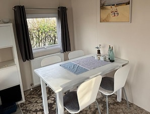 Una habitación con una mesa, sillas, un radiador, una ventana con cortinas y un cuadro enmarcado en la pared.