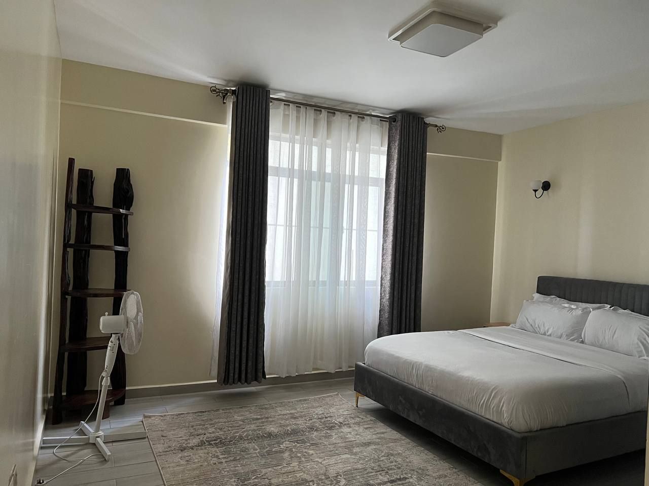 Un dormitorio con cama, ventilador, ventana con cortinas y lámpara de pared.