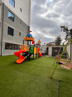 Un parque infantil con toboganes y columpios, frente a un edificio con ventanas amplias.