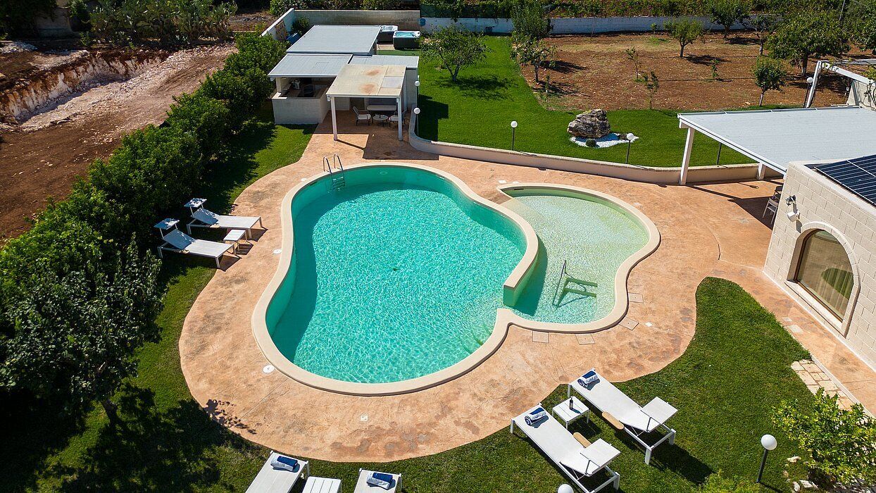 Piscina com borda curva, rodeada por um terraço com espreguiçadeiras e área de estar coberta.