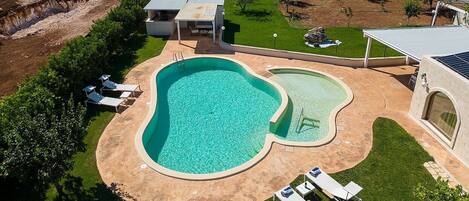 Ein Pool mit geschwungener Kante, umgeben von einer Terrassenfläche mit Liegestühlen und einem überdachten Sitzbereich.