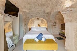A stone-walled room with a bed, a television mounted on the wall, and a bench with towels. - Villa Velar: A bright trullo in the characteristic style of the Apulian countryside, with Free WI-FI. (Carovigno)