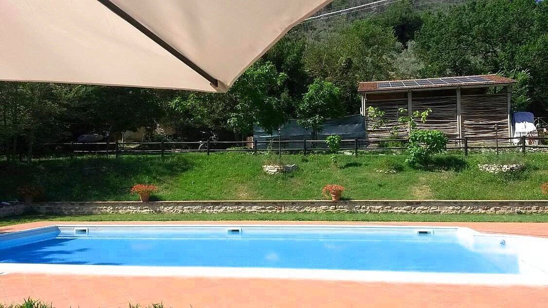 Une piscine dotée d’un espace ombragé, d’une zone délimitée par une clôture et, au fond, d’un bâtiment avec un toit en pente.