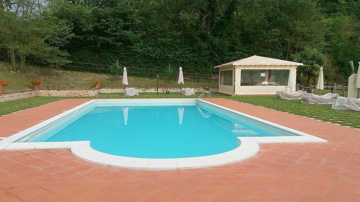 Piscina rectangular con solárium empedrado, rodeada de césped, árboles y un área de estar cubierta.