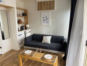 Een gezellig, compact ingerichte ruimte met een bank, een eettafel met twee borden en wijnglazen, een boekenkast en een lijst aan de muur.