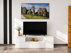 Un salon moderne avec un téléviseur à écran plat fixé sur un meuble blanc, un tableau représentant un paysage encadré au-dessus du téléviseur et une porte en bois sur la droite.