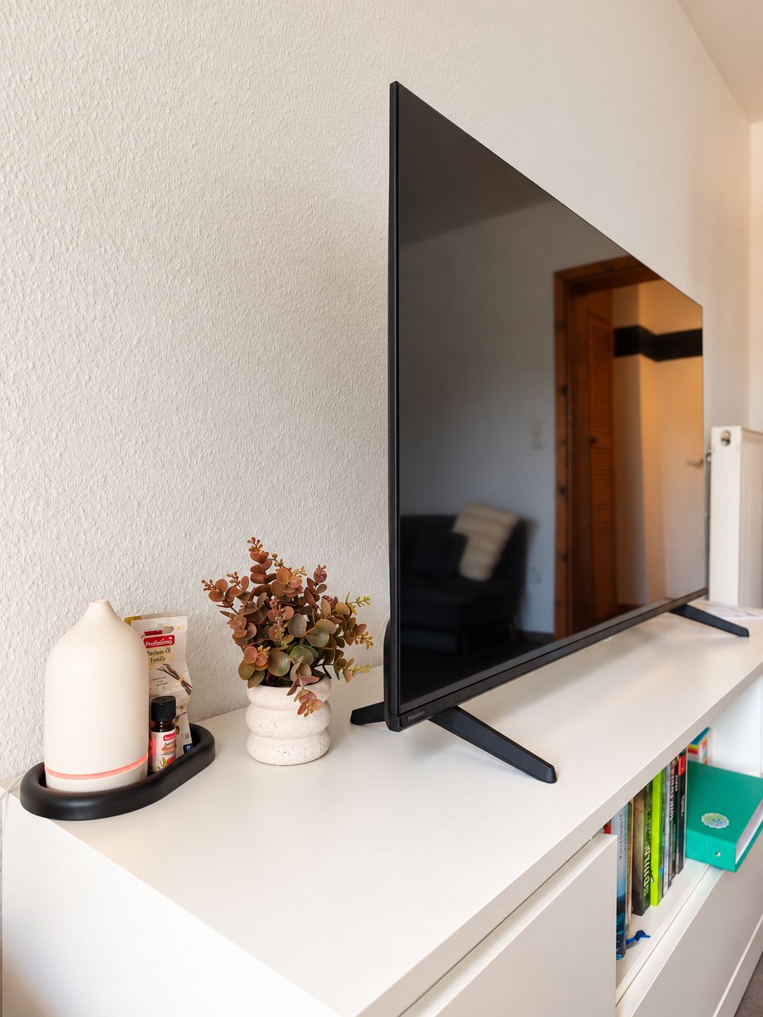 A flat-screen TV on a white shelf with a potted plant, a white vase, and a small container of coffee creamer.