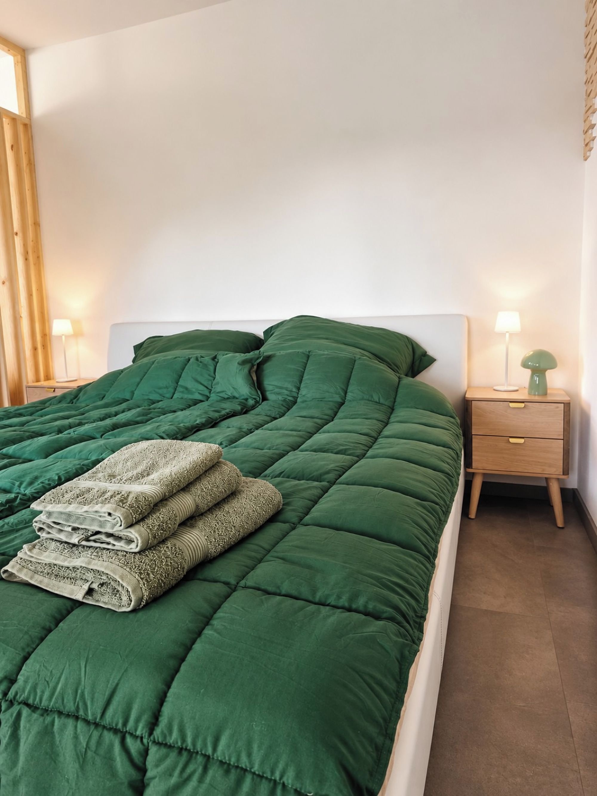Ein ordentlich bezogenes Bett mit einer grünen Bettdecke und Kissen, ein Nachttisch mit einer Lampe und eine Kommode.