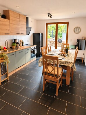 A modern kitchen with a dining area, featuring a large window, dark tiled flooring, and wooden furniture.