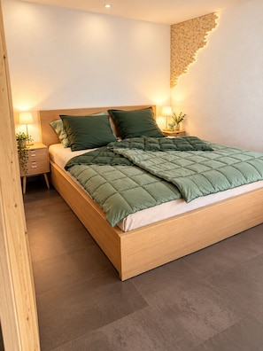 A modern bedroom with a wooden bed frame, green bedding, a nightstand with a lamp, and a wall-mounted light fixture.
