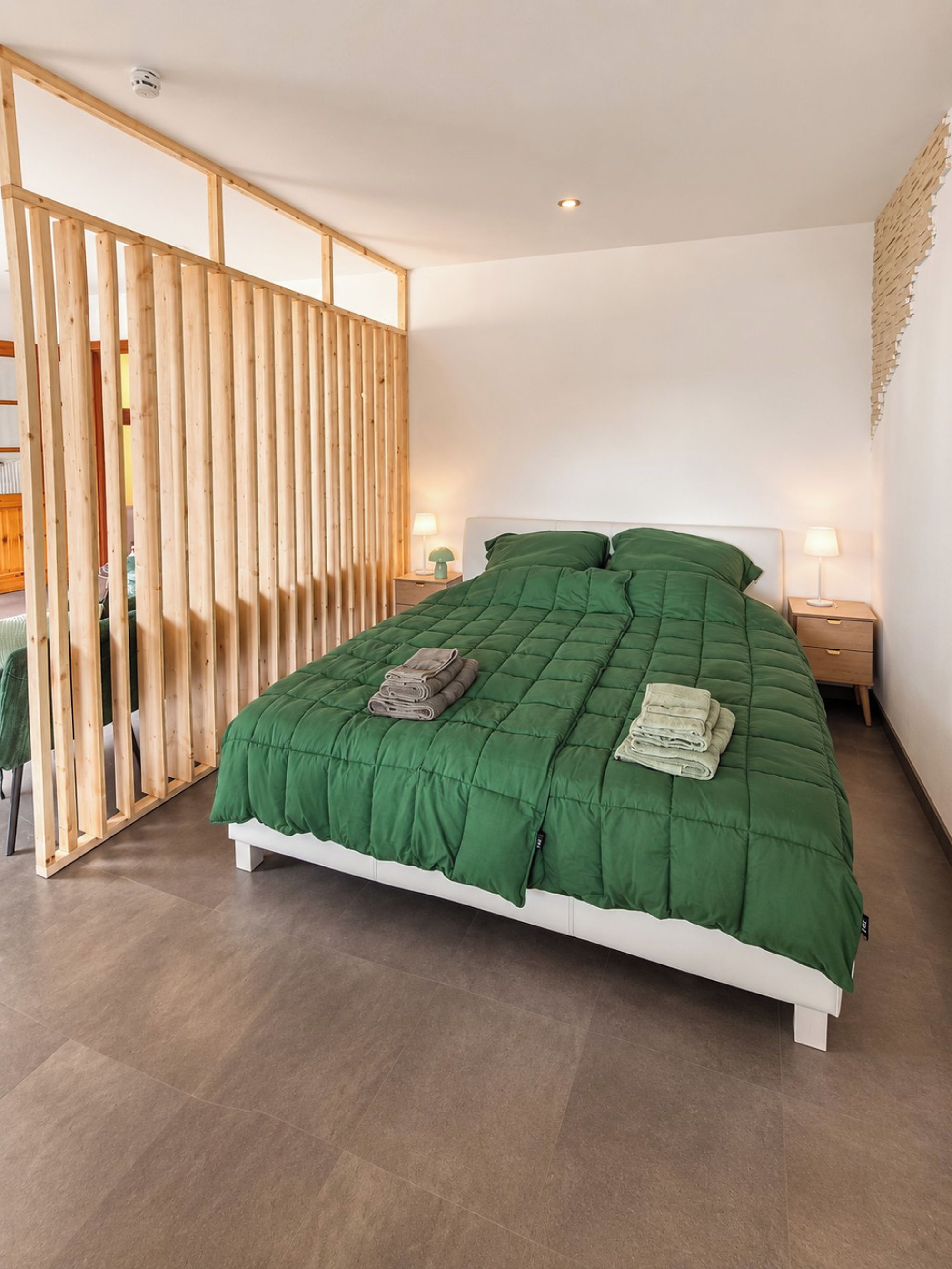 Ein Schlafzimmer mit einem großen Bett, Holzbalken, zwei Nachttischen mit Lampen und Blick auf einen Raum mit einem Heizkörper.