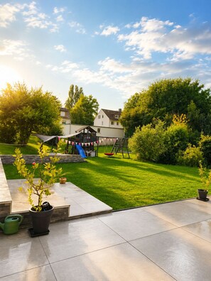 Un jardin avec un espace de jeux comprenant un toboggan et des balançoires, une terrasse agrémentée de plantes en pot et une pelouse.