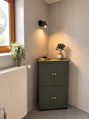 Ein Badezimmer im Eck mit Heizkörper, einem Schrank mit zwei Türen, einer Wandleuchte und einem Fenster mit einer Pflanze.