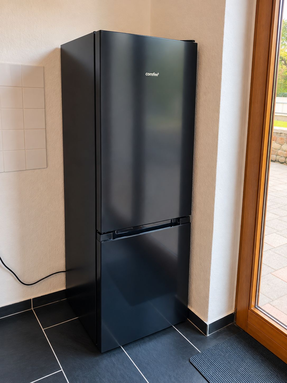 Ein schwarzer Kühlschrank der Marke 'comfee' steht in einer Ecke eines Raumes mit Fliesenboden und einem Fenster.